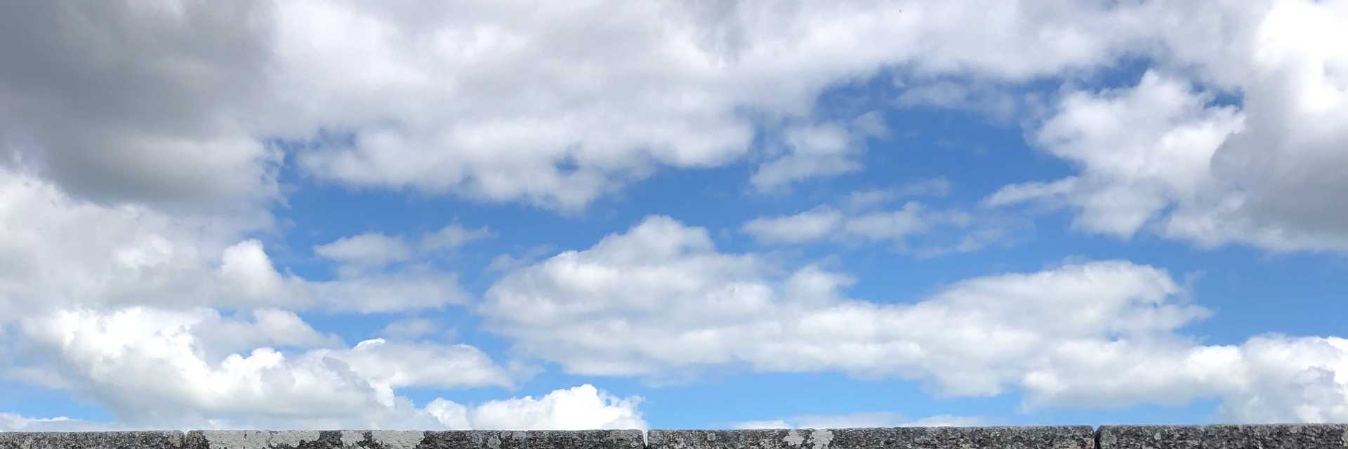 Photography of a stone wall running alongside a path. A bright blue sky and bright clouds are visible in the background.